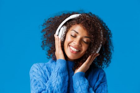 Close-up portrait happy smiling, romantic and tender african american woman enjoying listening music in headphones, tilt head close eyes dreamy and grinning delighted, blue background.