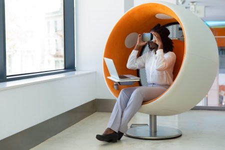 Female office employee with laptop watching virtual presentation. Black woman in office clothes and virtual reality goggles sitting in interactive chair and holding headset. VR experience concept